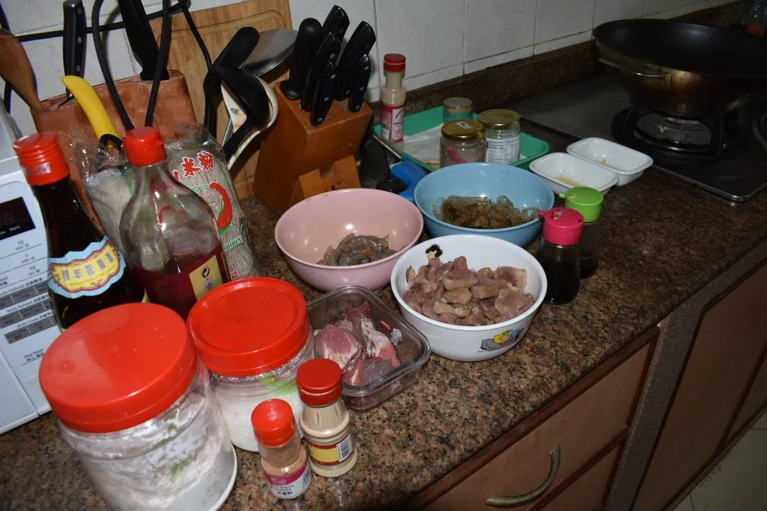 A kitchen counter covered in bowls of prepared prawns, pork and chicken beside sauces and a knife block.