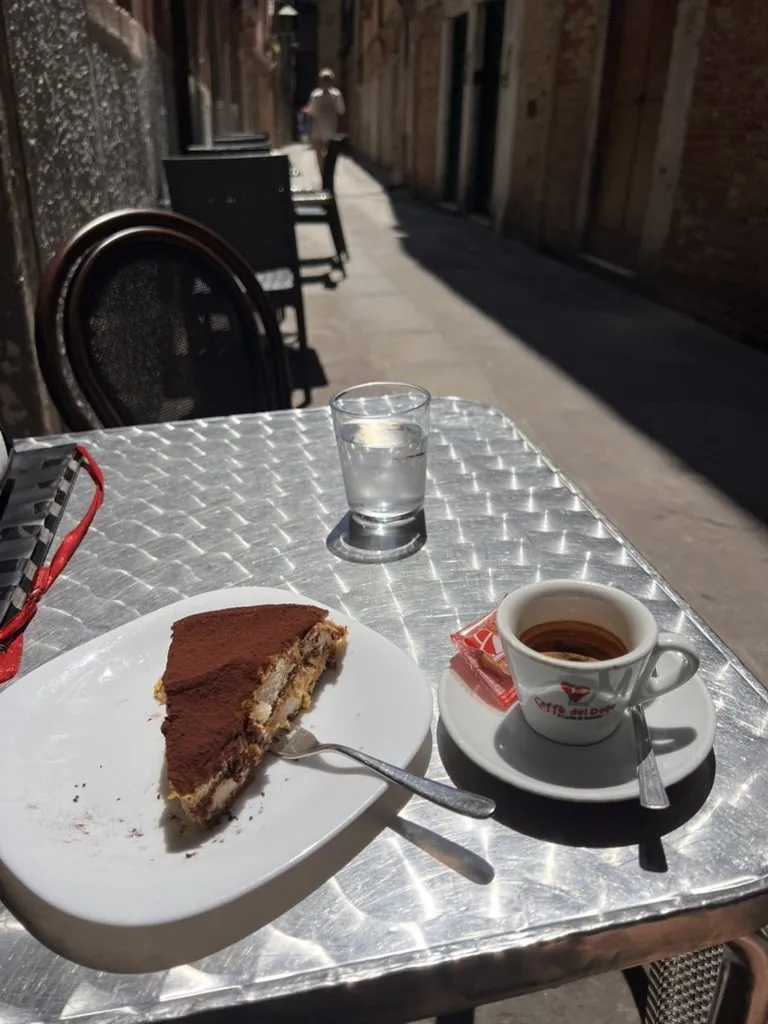 An espresso and a slice of tiramisu on a metal table in a narrow alley.